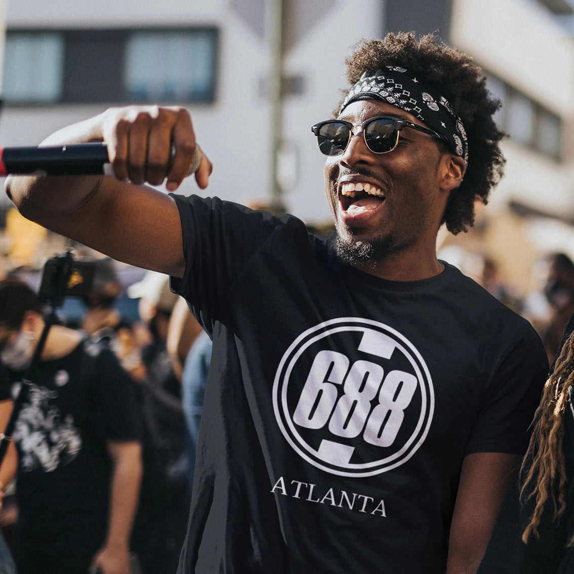 A man is wearing a black t-shirt with '688 Atlanta' logo, holding a microphone, in an urban setting.