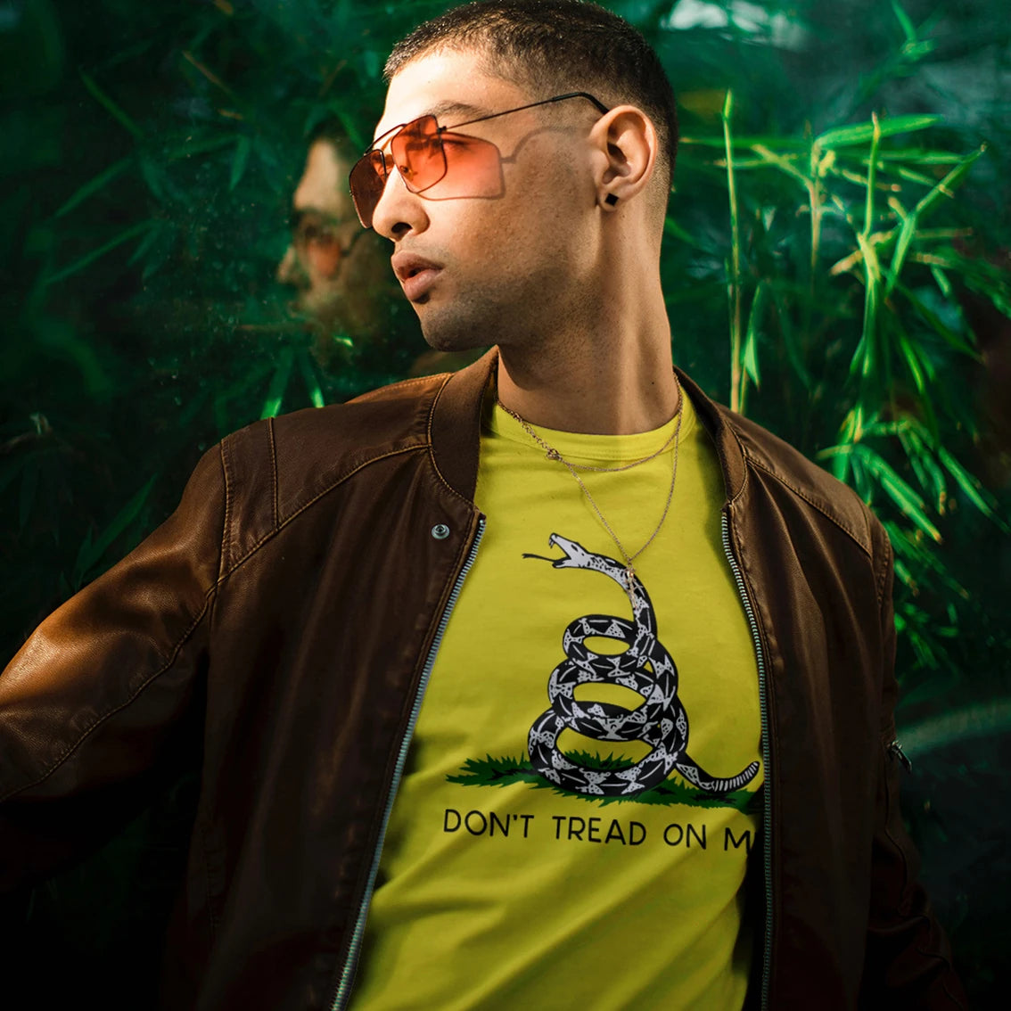 A man in sunglasses is wearing a yellow t-shirt with a snake graphic and 'DON'T TREAD ON ME' text, standing against a green leafy background.