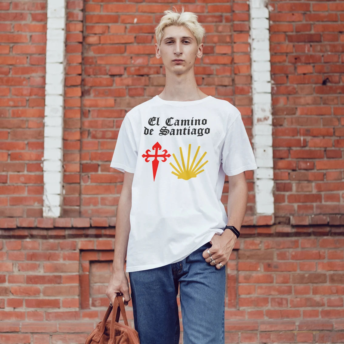 A man is wearing a white t-shirt with 'El Camino de Santiago' design against a brick wall.
