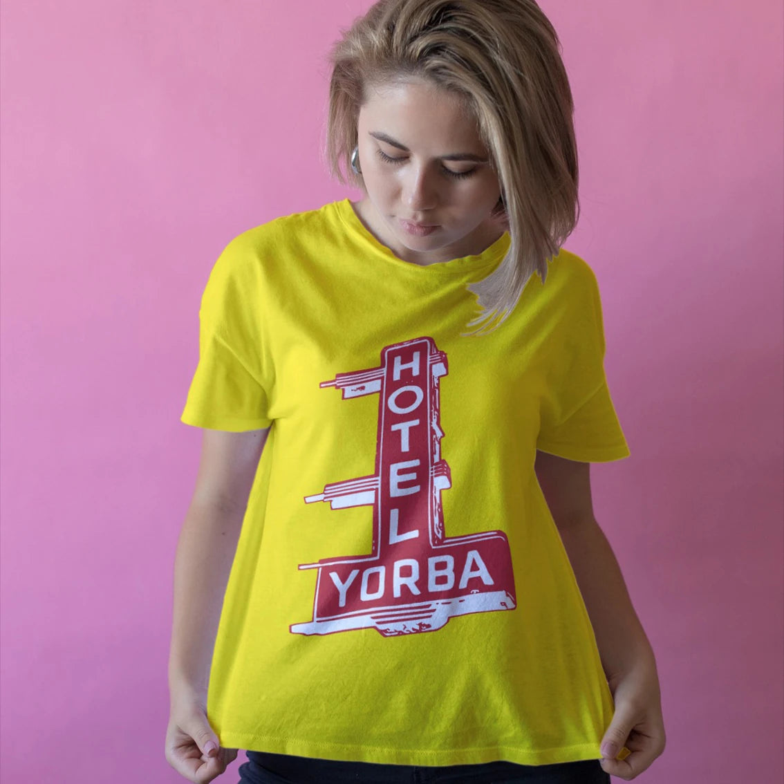 A woman is wearing a yellow t-shirt with 'HOTEL YORBA' sign design against a pink background