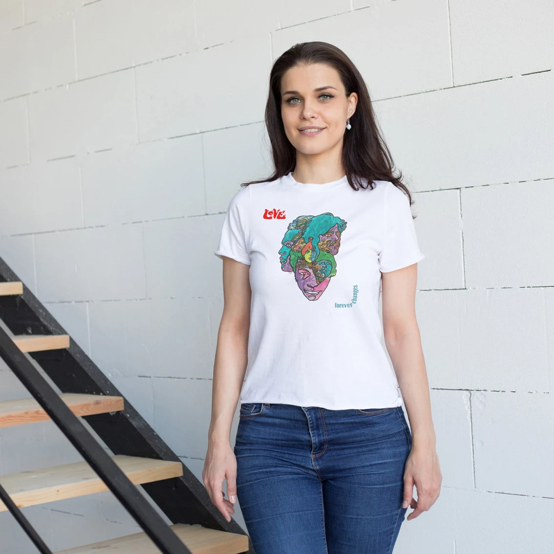 A woman is wearing a white t-shirt with a colorful graphic design with text on the left side saying 'Love' and 'Forever Changes' on the right against a light brick wall.