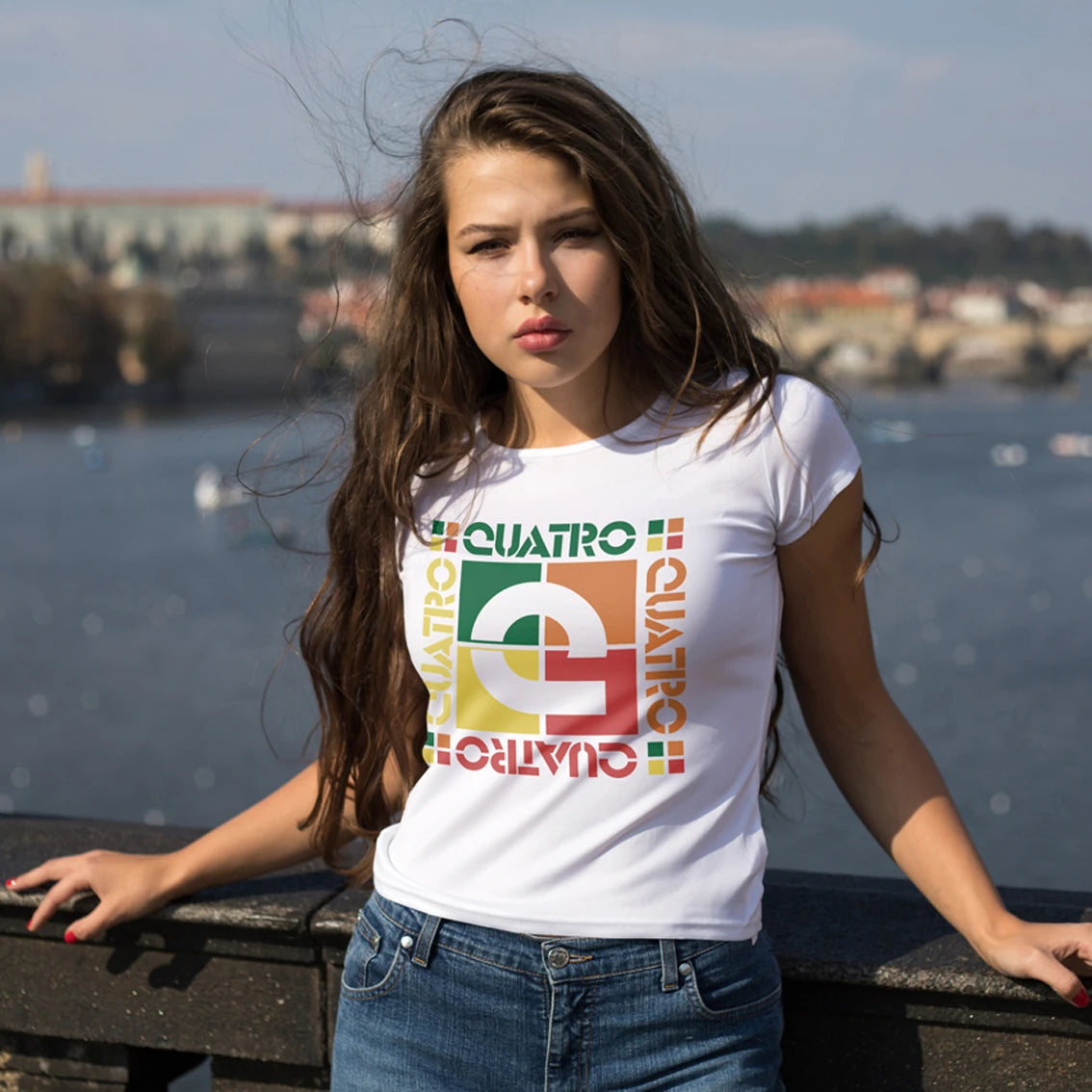 A woman is wearing a white t-shirt with colorful 'CUATRO' logo by a waterfront.