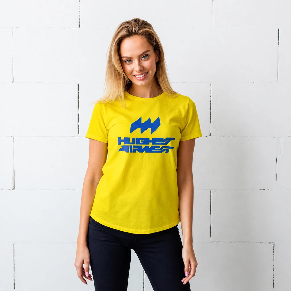 A woman wearing a bright yellow t-shirt with a blue 'HUGHES AIRWEST' logo on the chest, standing against a white painted brick wall.