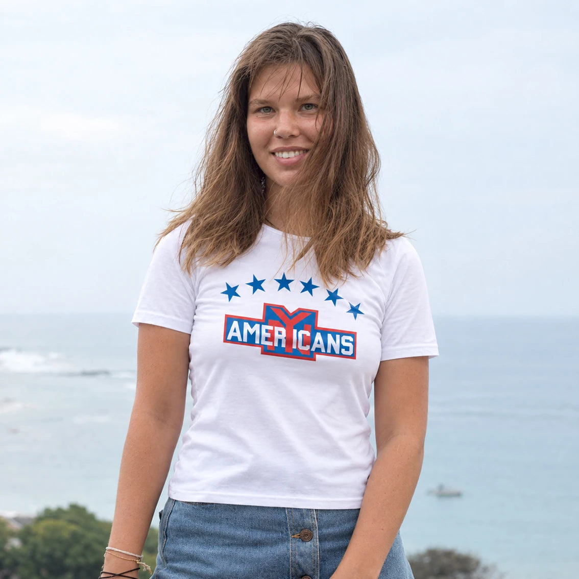 A woman wearing a white graphic t-shirt with 'NY AMERICANS' design in red, white, and blue, standing outdoors with an ocean background.