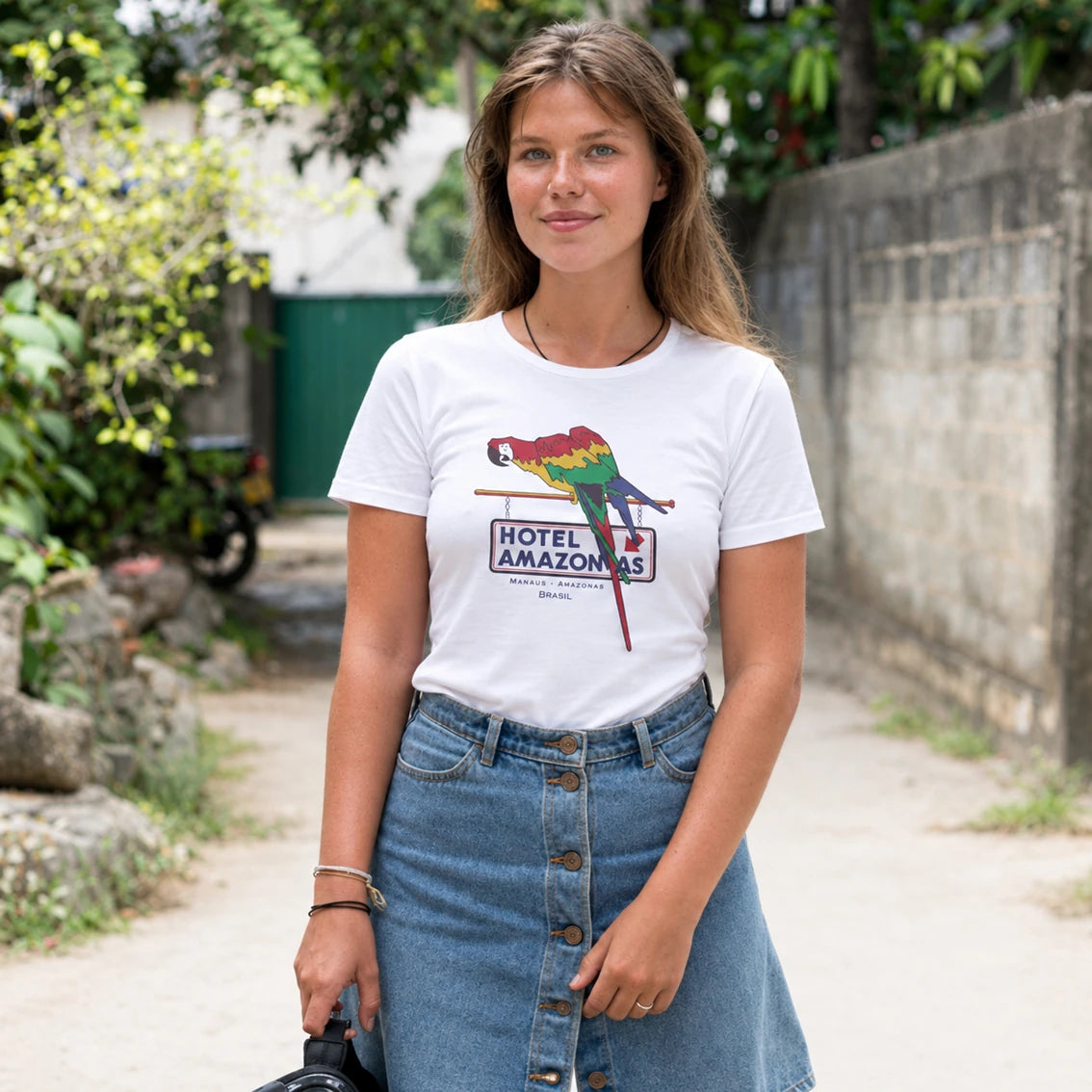 A woman wearing a white graphic t-shirt with a colorful parrot and 'Hotel Amazonas' print, paired with a blue denim skirt, standing outdoors on a path with green foliage and a stone wall in the background.