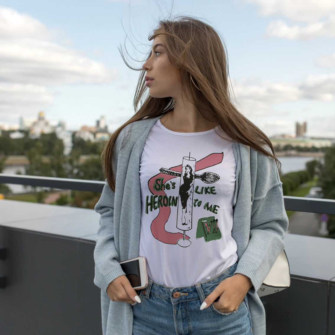 Woman wearing a graphic t-shirt with 'She's Like Heroin To Me' text and design, standing on a rooftop with a cityscape background.