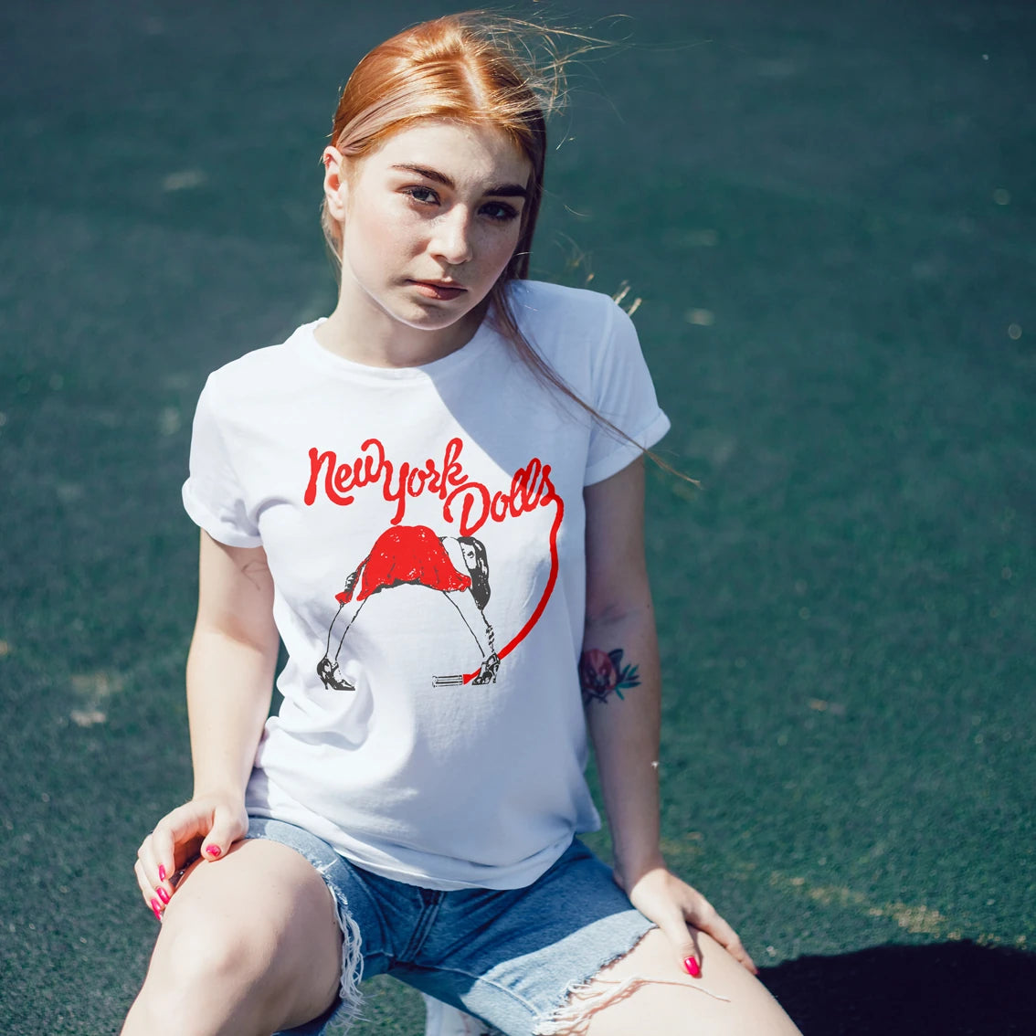 Woman wearing a white t-shirt with 'New York Dolls' graphic design, sitting on a grassy area.