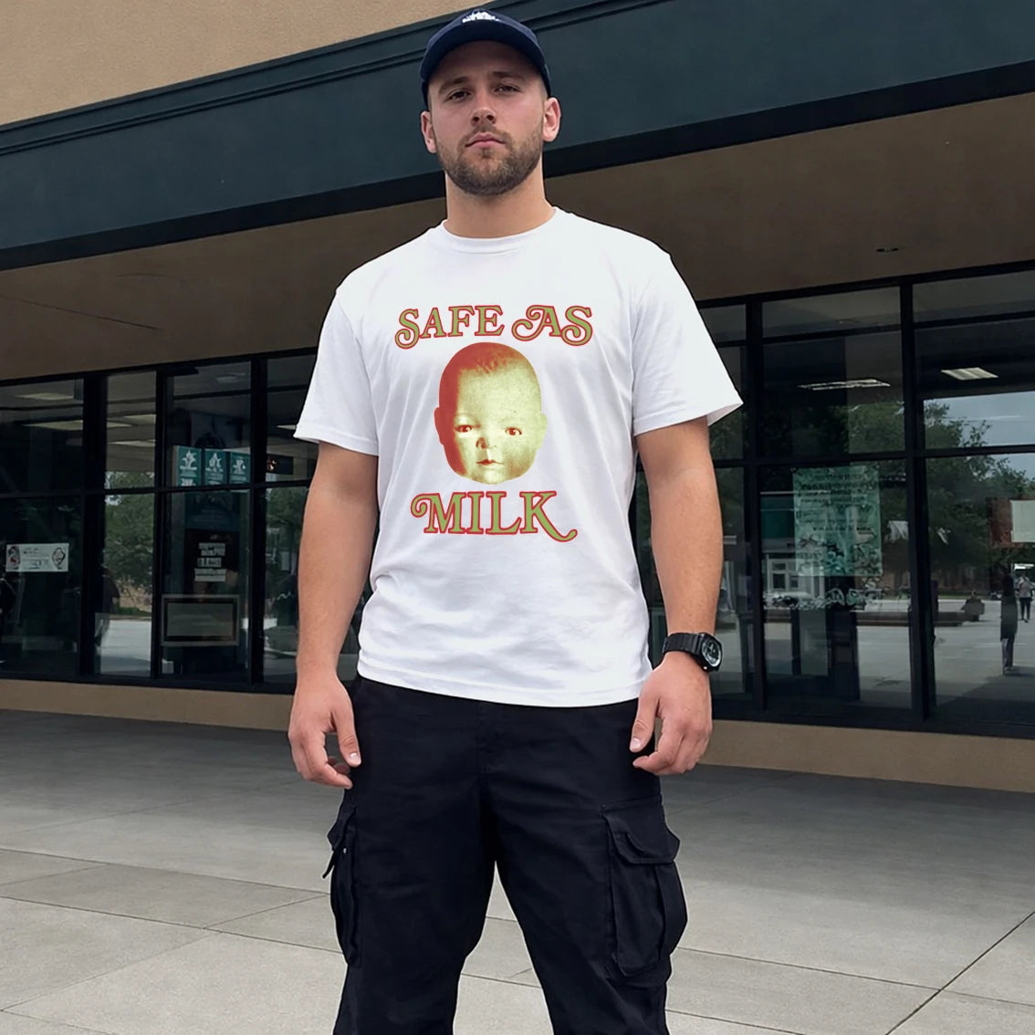 Man in hat wearing a white t-shirt with a baby face graphic design and 'Safe As Milk' text, standing outside a building.