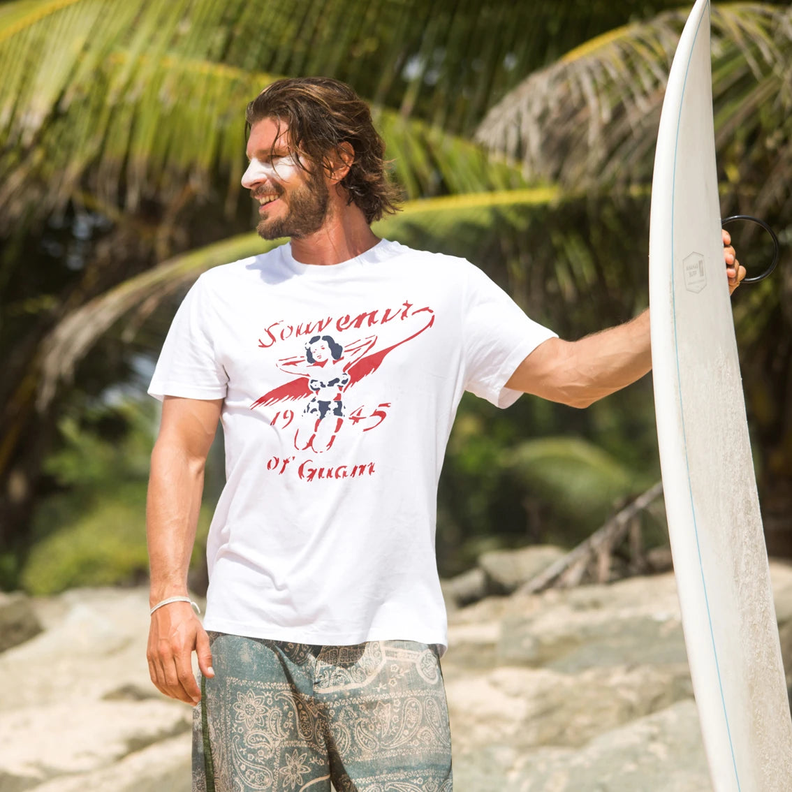 Man holding a surfboard and wearing t-shirt with 'Souvenir Of Guam -1945' text with a tropical background