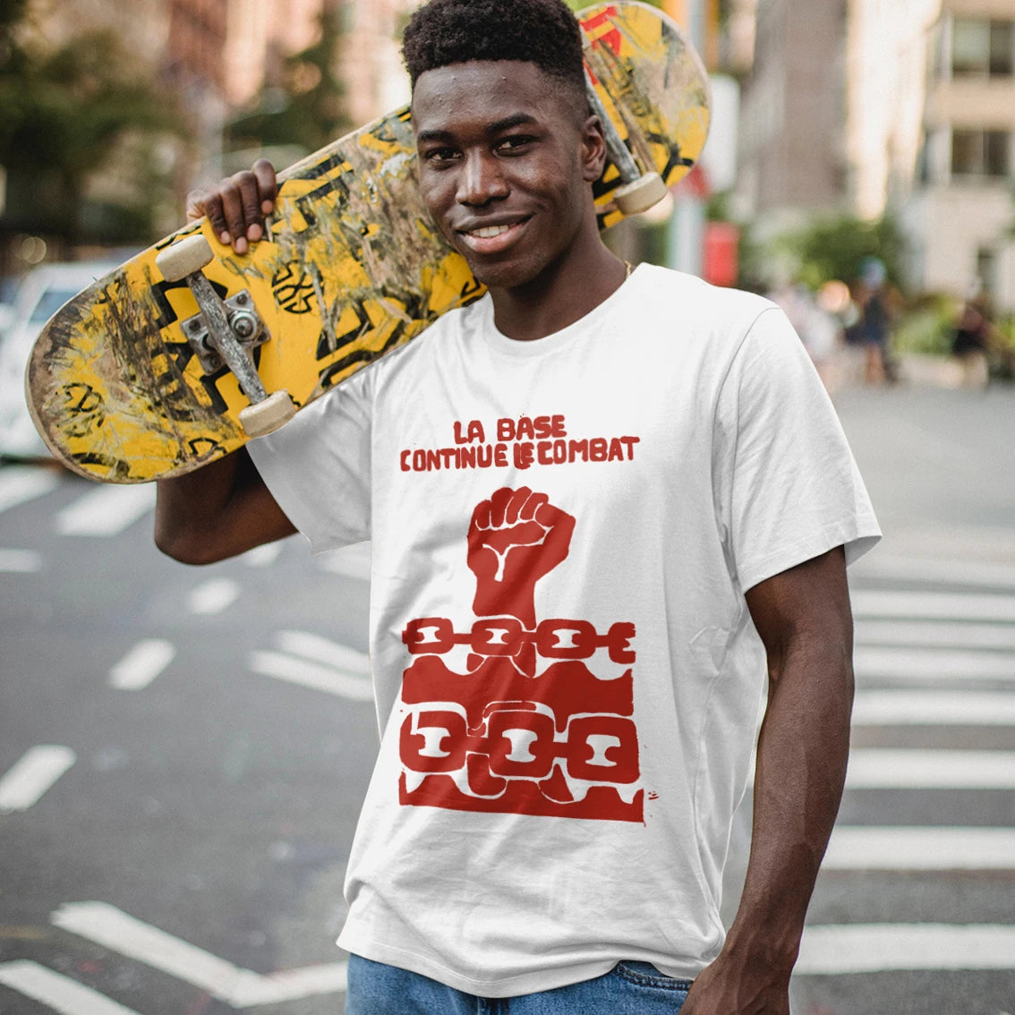 Man wearing a white t-shirt with red graphic design and 'La Base Continue Le Combat' text, holding a skateboard on a city street.