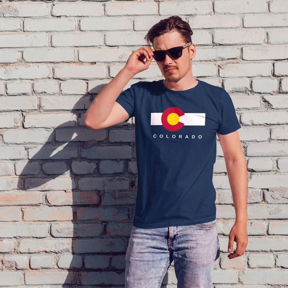 Man in sunglasses wearing a navy blue t-shirt with a Colorado flag design against a light brick wall.
