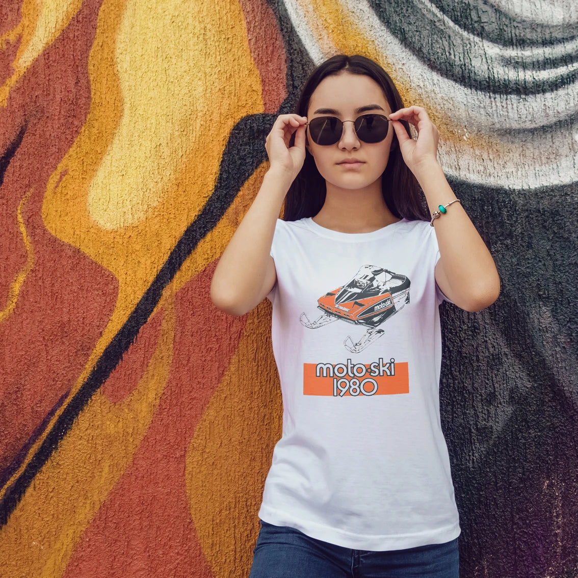 Woman wearing a white t-shirt with a graphic design of a racing car and text 'Moto Ski 1980' in front of a colorful wall.