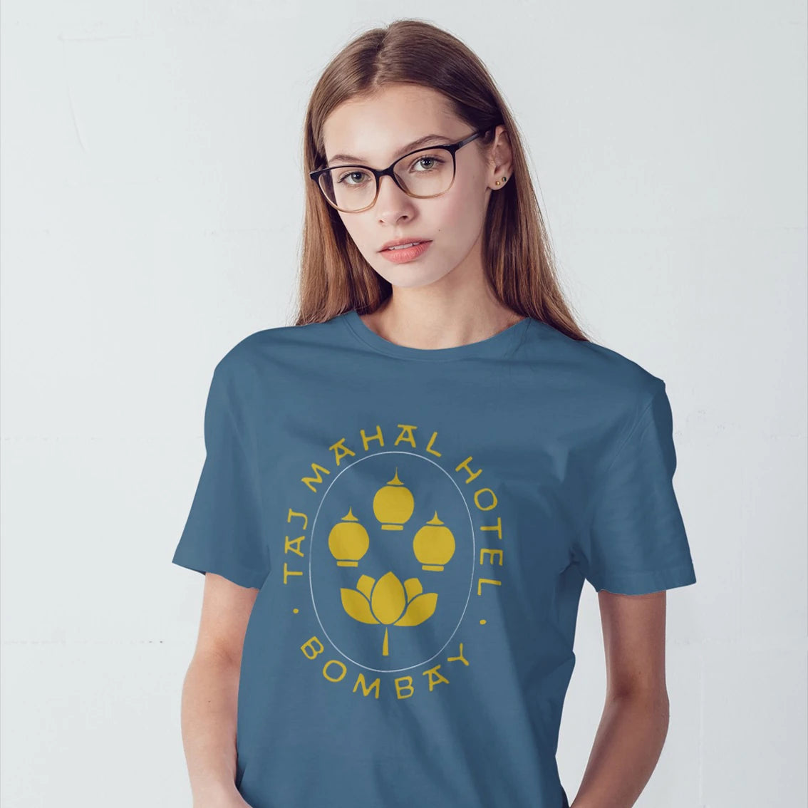 A woman in glasses is wearing a blue t-shirt with 'Taj Mahal Hotel, Bombay' design on a white background