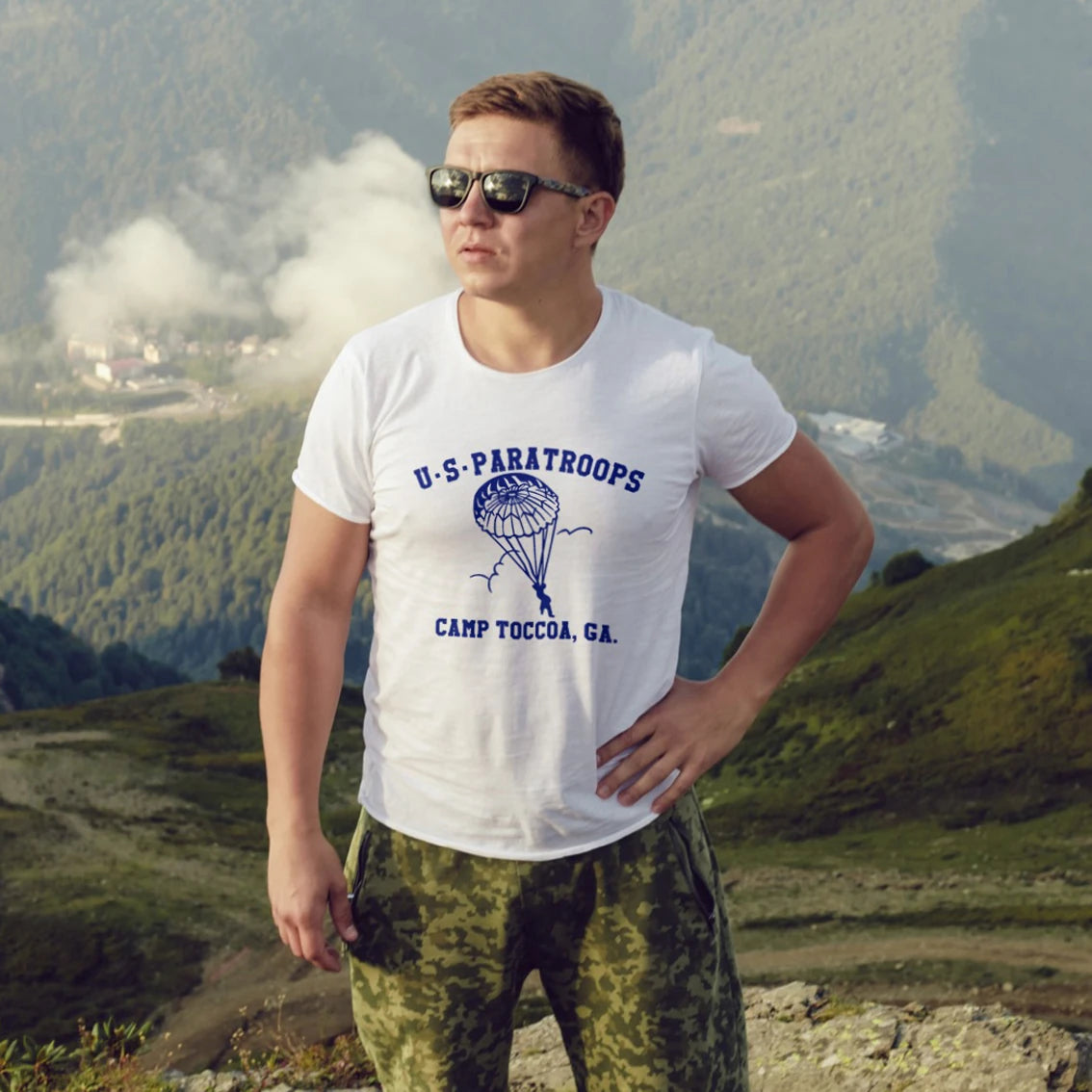 A man in sunglasses is wearing a white t-shirt with 'U.S. Paratroops Camp Toccoa, GA' design in a mountainous landscape