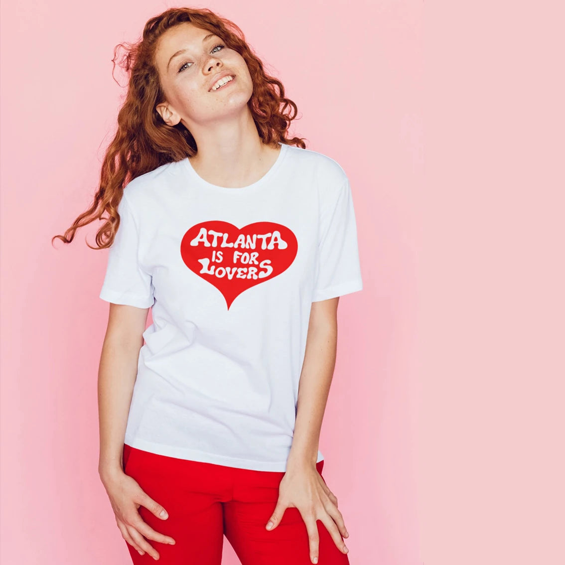 A woman is wearing a white t-shirt with white letters saying 'Atlanta is for Lovers' on a red heart shape design on a pink background