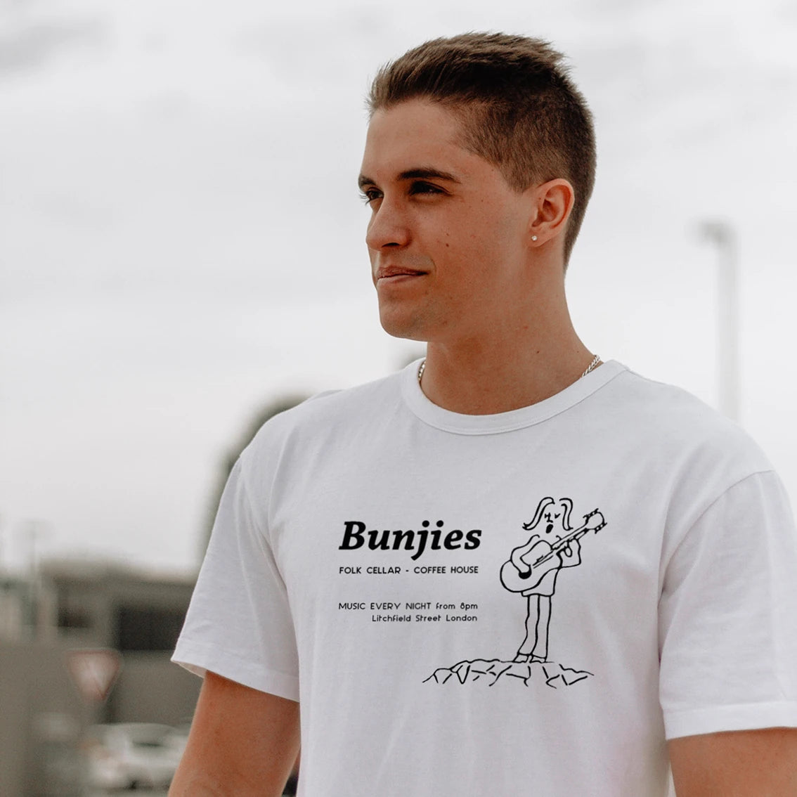 A man is wearing a white t-shirt with 'Bunjies - Folk Cellar - Coffee House' text and graphic design of a bunny with a guitar, in built up area