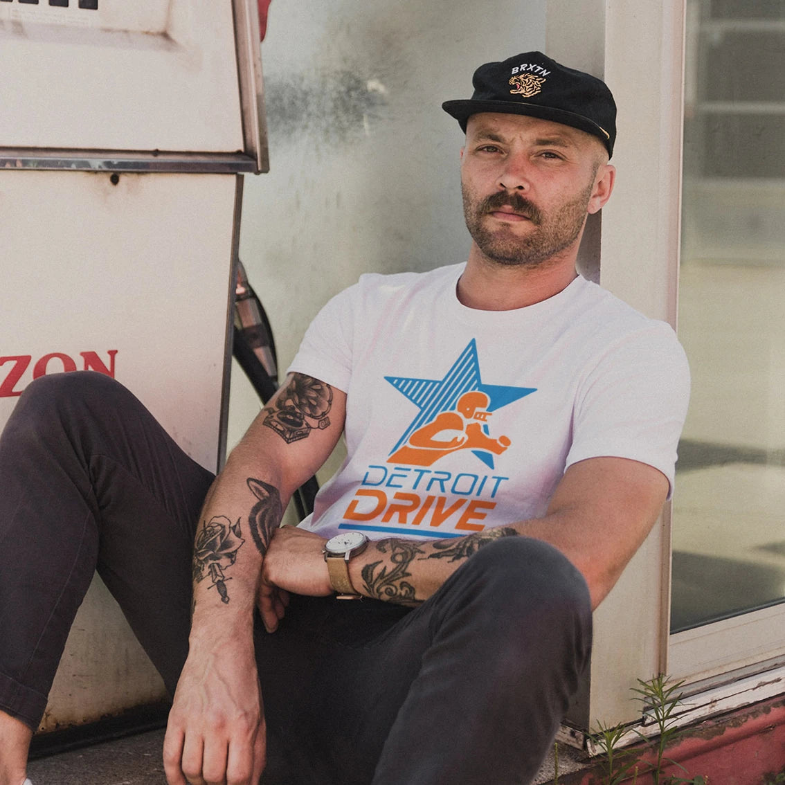 A man is wearing a 'Detroit Drive' t-shirt with a graphic design of a star and figure holding a football on it, sitting next to a house.