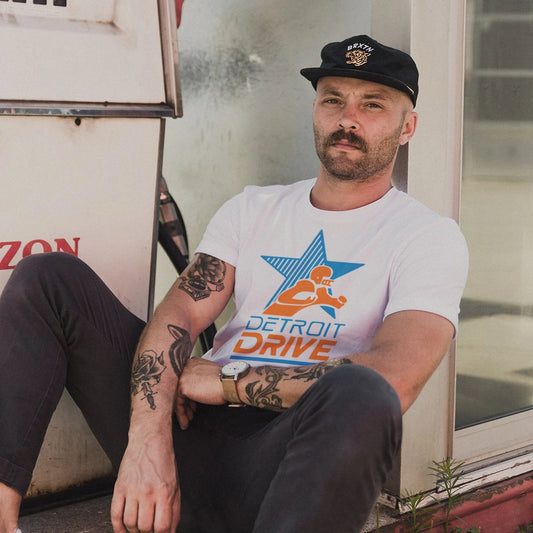 A man is wearing a 'Detroit Drive' t-shirt with a graphic design of a star and figure holding a football on it, sitting next to a house.
