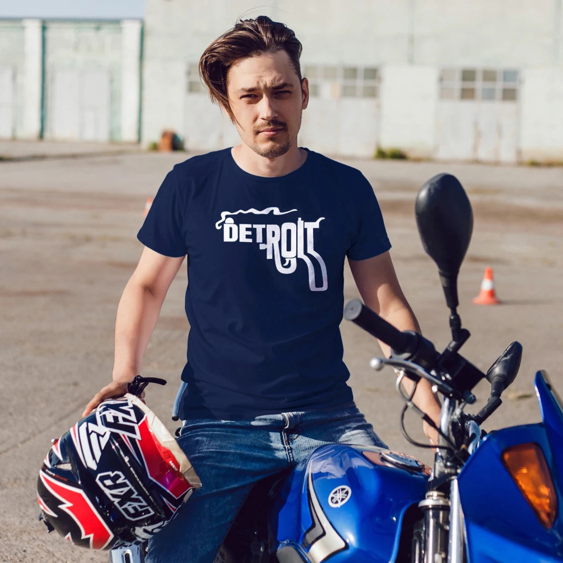 A man is wearing a navy blue t-shirt with 'Detroit' text and smoking gun design, sitting on a blue motorcycle.
