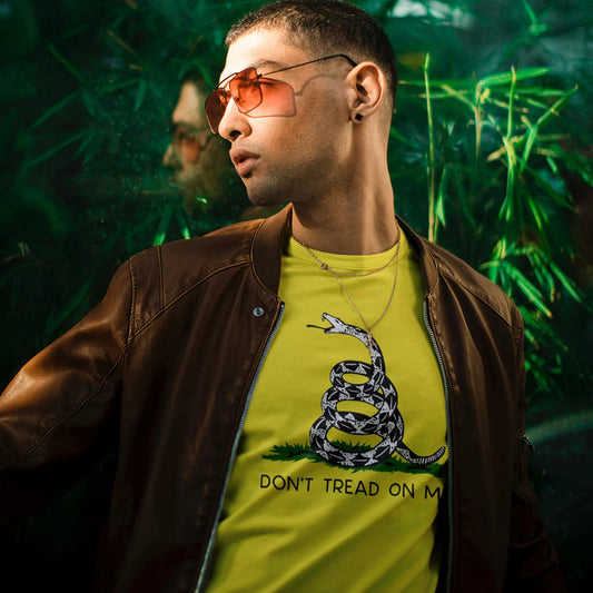 A man in sunglasses is wearing a yellow t-shirt with a snake graphic and 'DON'T TREAD ON ME' text, standing against a green leafy background.