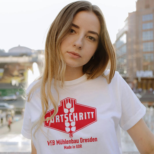 A woman is wearing a white t-shirt with a red logo and 'FORTSCHRITT - VEB Muhlenbau Dresden, Made in GDR' text, standing outdoors.