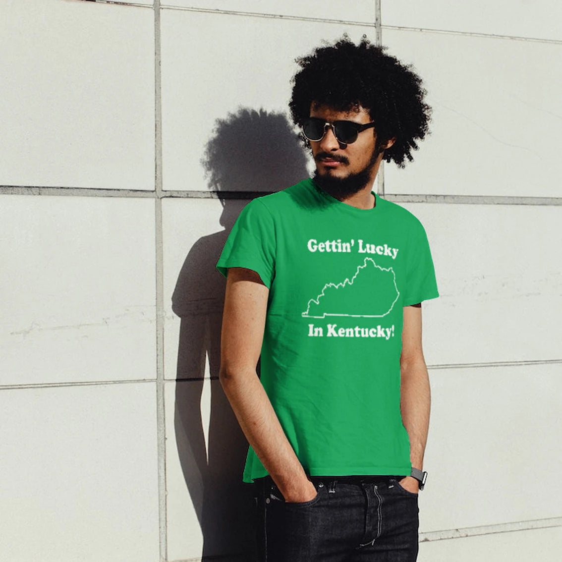 A man in sunglasses is wearing a green t-shirt with 'GETTIN' LUCKY IN KENTUCKY' text and a map, standing against a light-colored wall.
