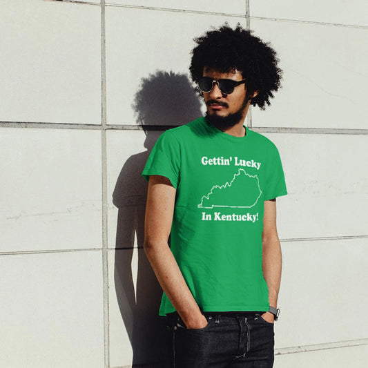 A man in sunglasses is wearing a green t-shirt with 'GETTIN' LUCKY IN KENTUCKY' text and a map, standing against a light-colored wall.