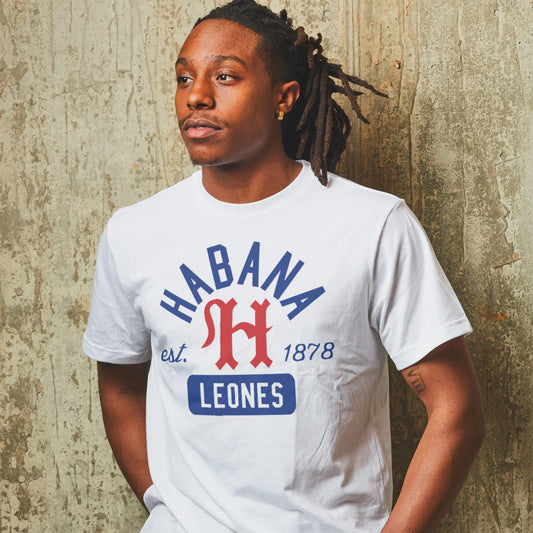 A man is wearing a white t-shirt with 'Habana Leones -est. 1878' text and letter H logo design against a textured wall.