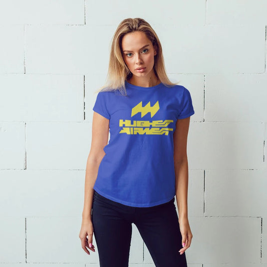 A woman is wearing a blue t-shirt with a yellow logo and 'HUGHES AIRWEST' text against a light gray brick wall.