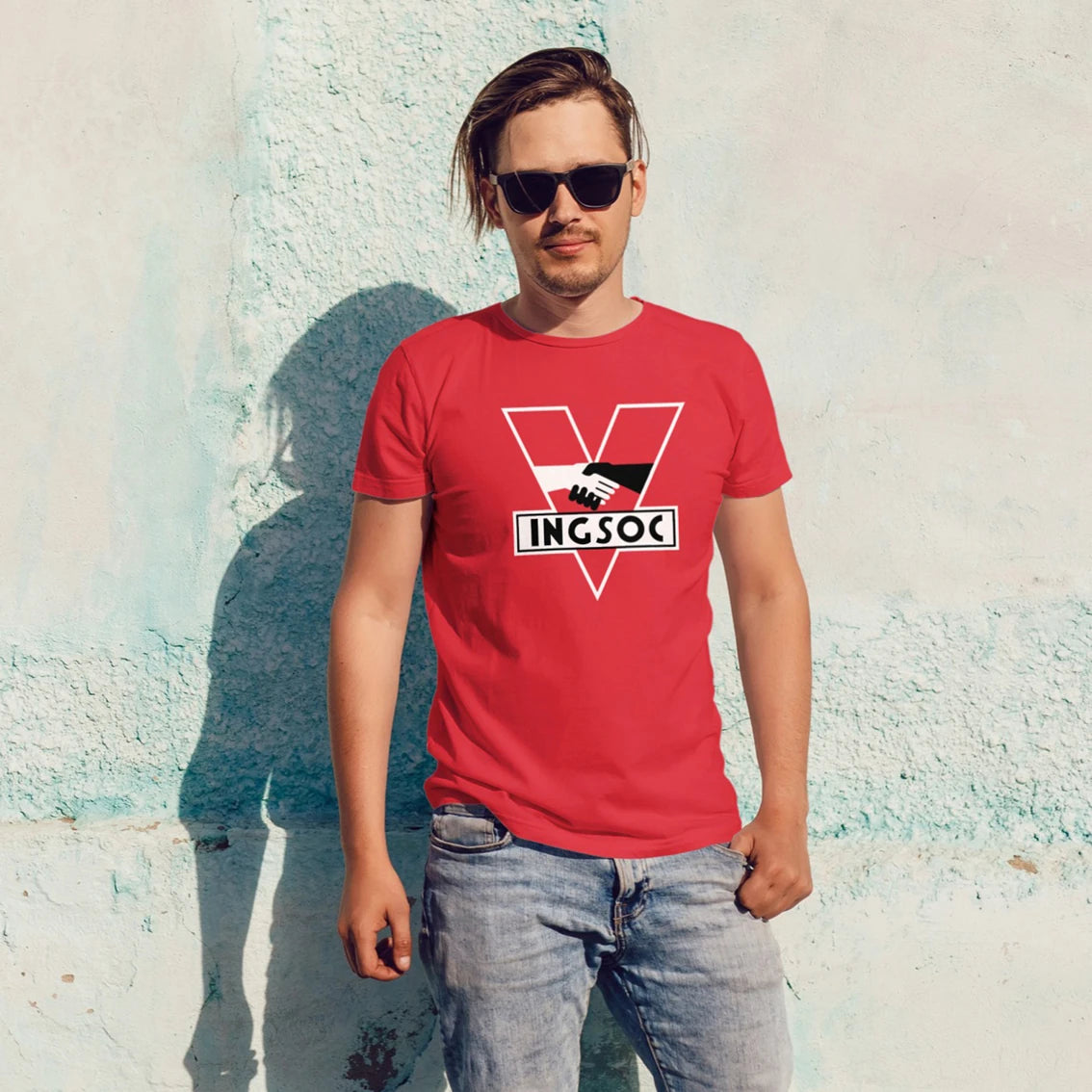 A man in sunglasses is wearing a red t-shirt with a V-shape and handshake logo and 'INGSOC' text against a textured wall.