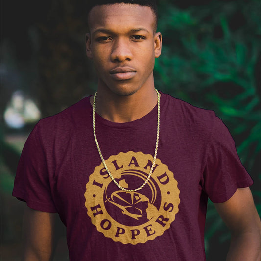 A man is wearing a metal necklace and maroon t-shirt with a round shape logo and 'ISLAND HOPPERS' text, standing against a blurred green background