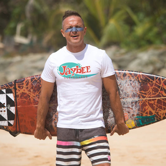 A man is holding a surfboard on a beach wearing a white t-shirt with 'Jaybee Surfboards' branding.