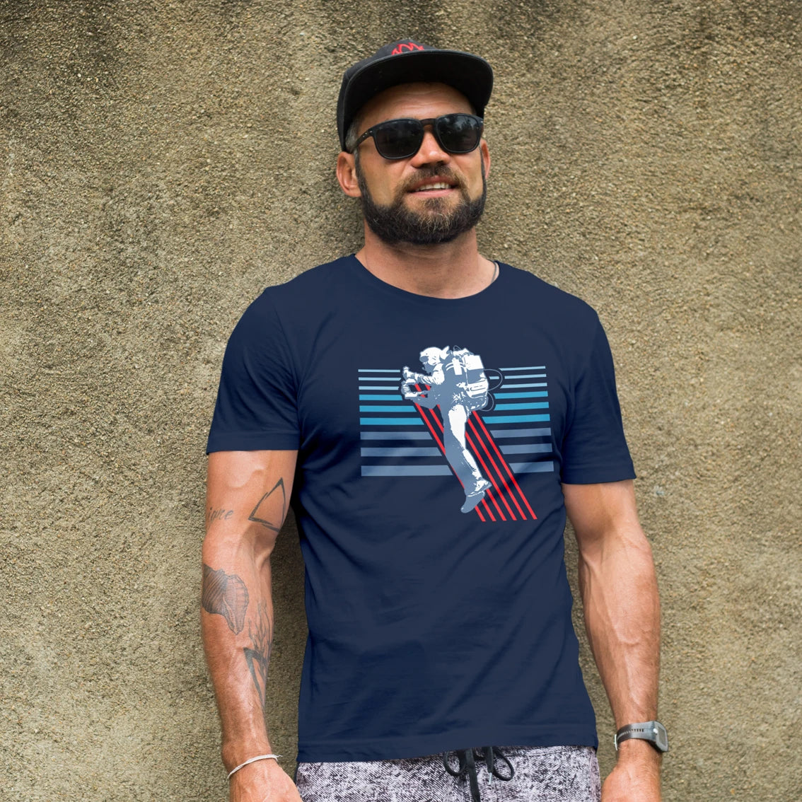 A man in sunglasses and hat is wearing a navy blue t-shirt with a graphic design of a human with a jet pack on it against a textured wall.
