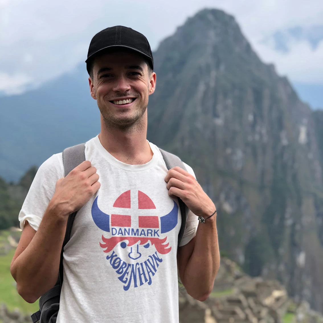 A man in a hat is wearing a t-shirt with 'Danmark København' text and viking hat in a shape of danish flag on it with mountains in the background