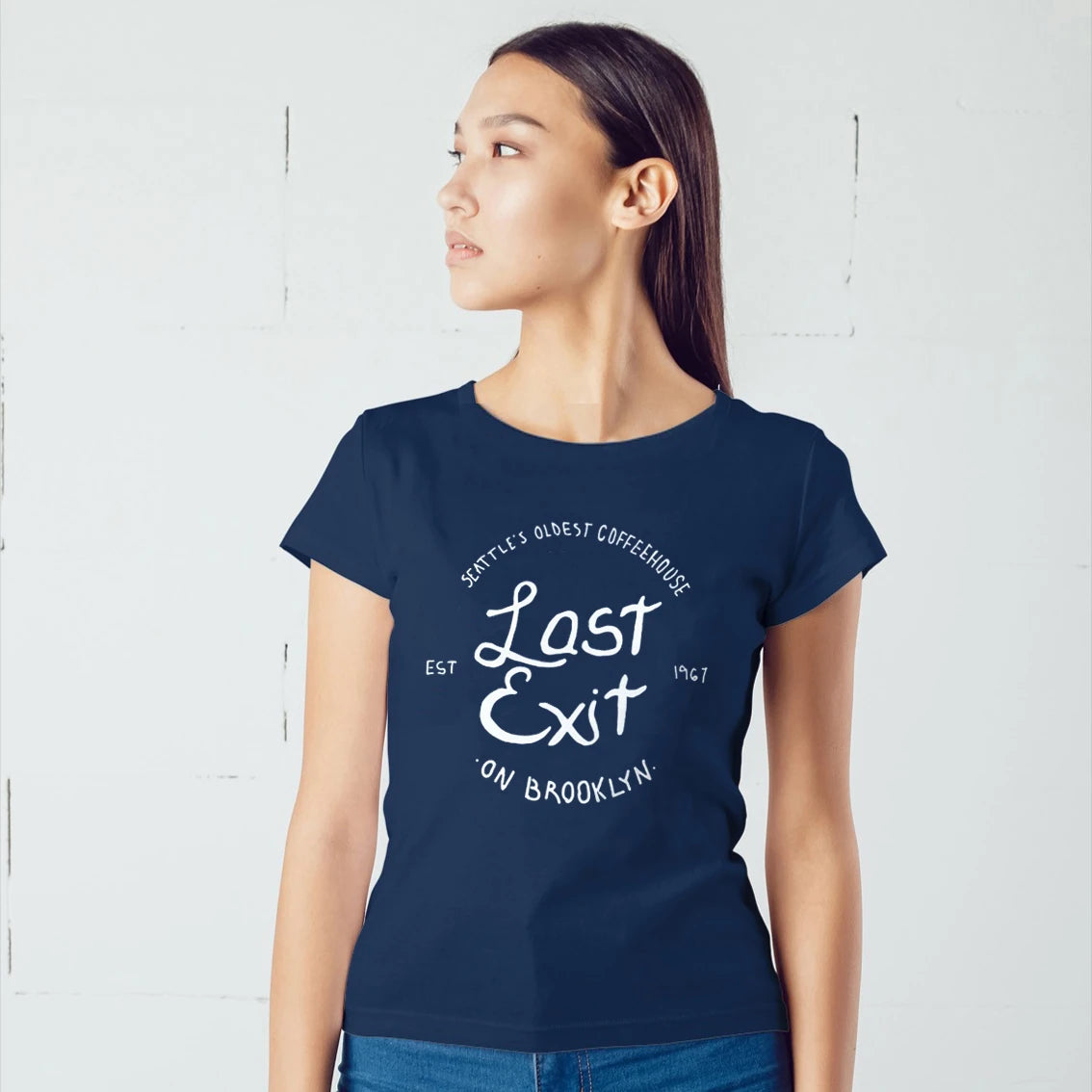 A woman is wearing a navy blue t-shirt with white 'Seattle's Oldest Coffeehouse - LAST EXIT ON BROOKLYN' text against a light gray background