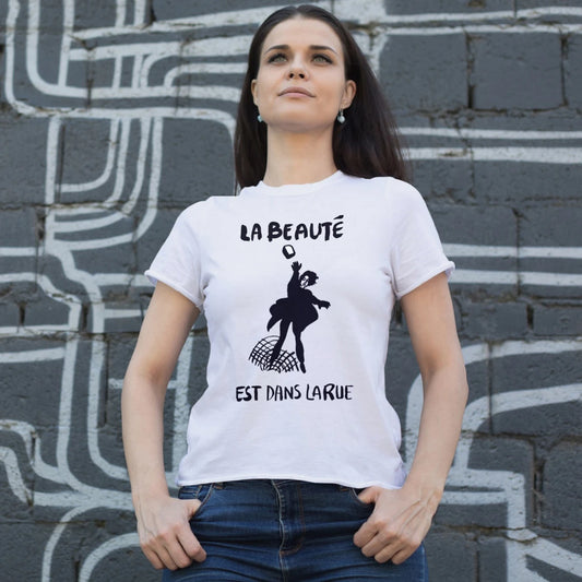 A woman is wearing a white t-shirt with a picture of the woman on it throwing a brick and black graphic text saying 'La Beaute Est Dans Larue'  against a grey wall.