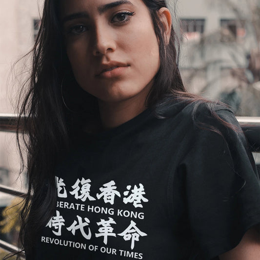 A woman is wearing a black t-shirt with a 'LIBERATE HONG KONG, REVOLUTION OF YOUR TIMES' white text in Chinese and English, standing on the balcony.