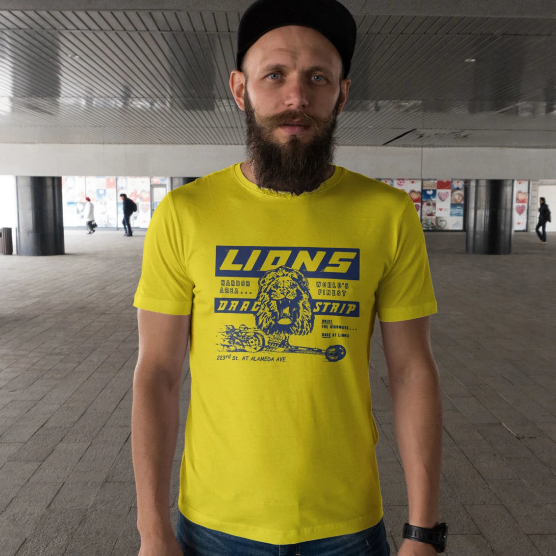 A man in a hat is wearing a bright yellow t-shirt with 'Lions Drag Strip' text and graphic design of the lion on it in an urban setting.