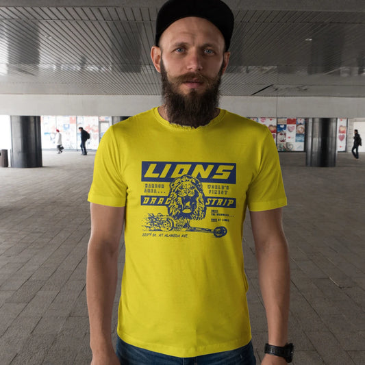 A man in a hat is wearing a bright yellow t-shirt with 'Lions Drag Strip' text and graphic design of the lion on it in an urban setting.
