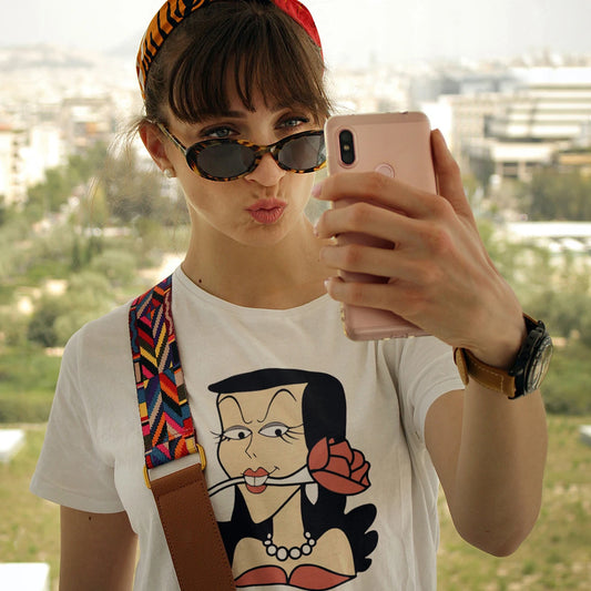 A woman in sunglasses is taking a selfie outdoors wearing a white t-shirt with a cartoon character design Natasha Fatale holding a rose in her mouth.