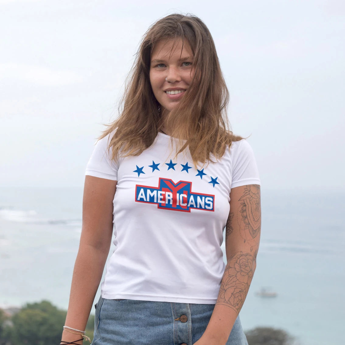 A woman is wearing a white t-shirt with design of the stars and  'AMERICANS' text, standing outdoors with the sea view