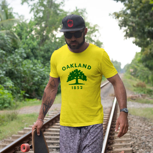 A man in a hat is wearing a yellow 'Oakland 1852' t-shirt on train tracks with greenery in the background