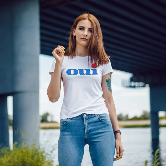 A woman is wearing a white t-shirt with 'OUI' printed on it, standing outdoors by a body of water.