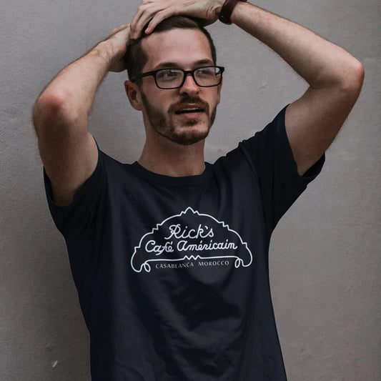 A man in glasses is wearing a black t-shirt with a logo and 'RICK'S CAFÉ AMÉRICAIN, Casablanca Morocco' text against a grey background