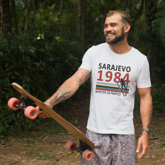 A man holding a skateboard is wearing white t-shirt with a 'Sarajevo 1984 Winter Olympics' logo in a forest setting