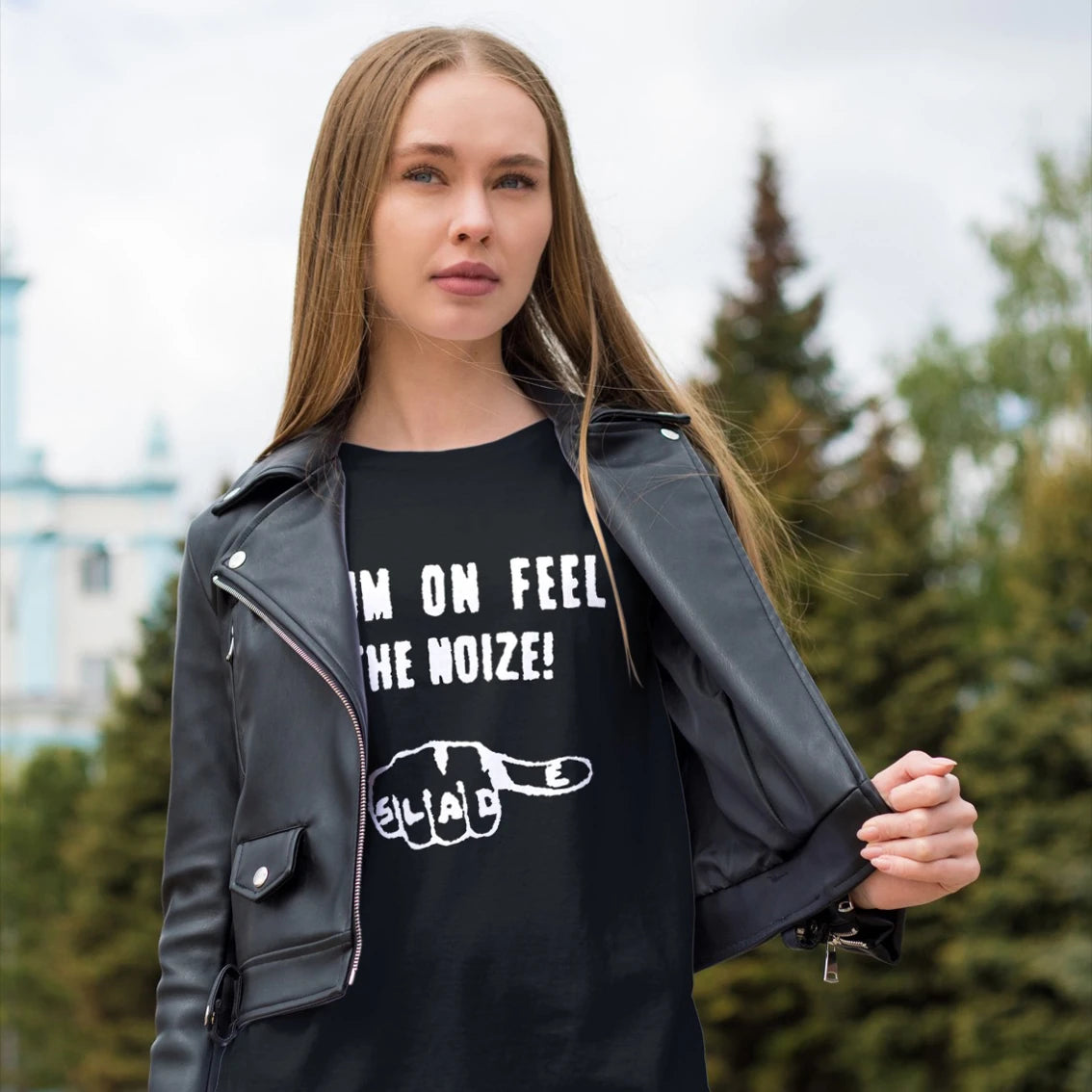 A woman is wearing a black leather jacket and black t-shirt with text 'SLADE CUM ON FEEL THE NOIZE' and picture of a fist on it outdoors