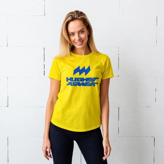A woman wearing a bright yellow t-shirt with a blue 'HUGHES AIRWEST' logo on the chest, standing against a white painted brick wall.