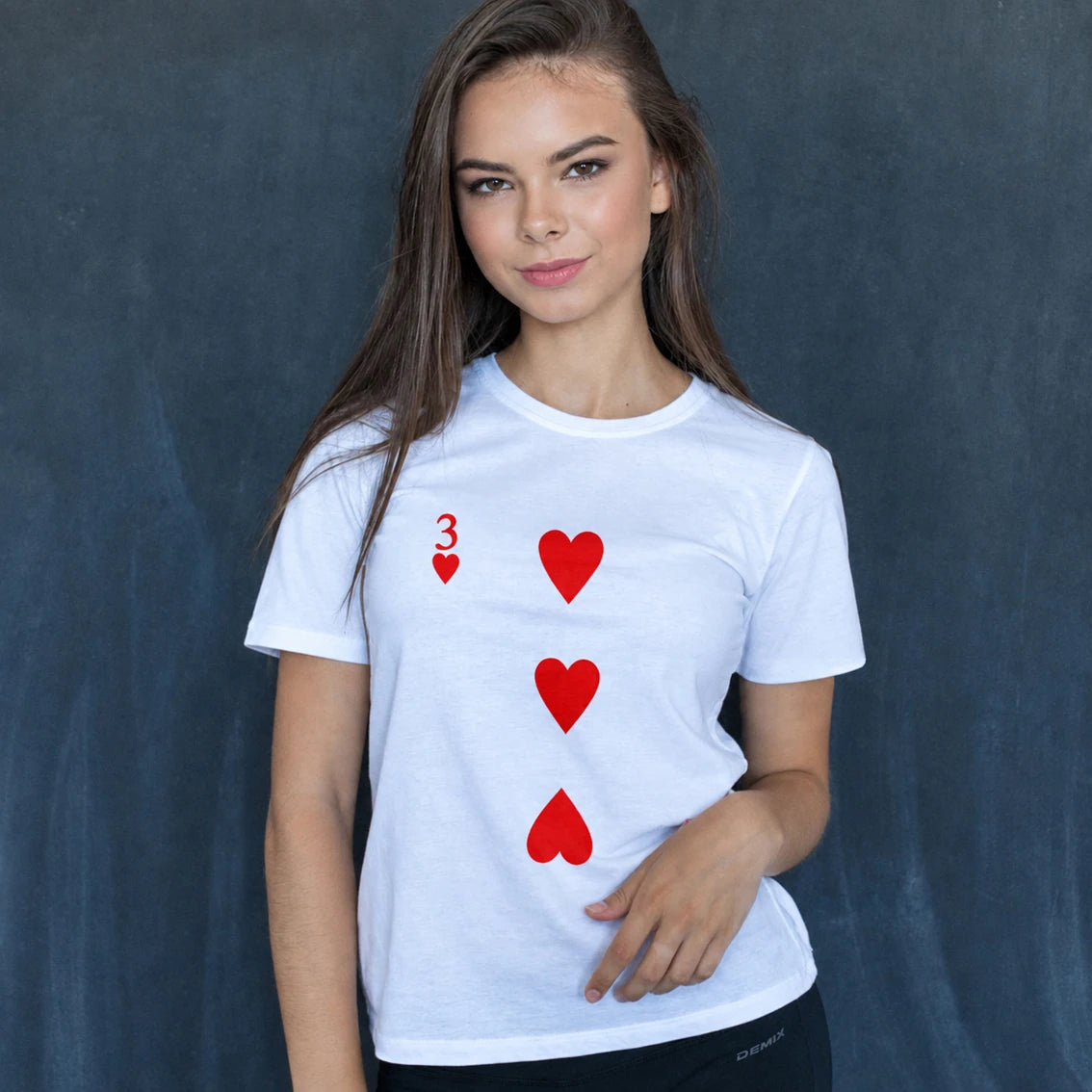 A woman modeling a white short-sleeve t-shirt with a red playing card design of the three of hearts, set against a dark textured studio backdrop.