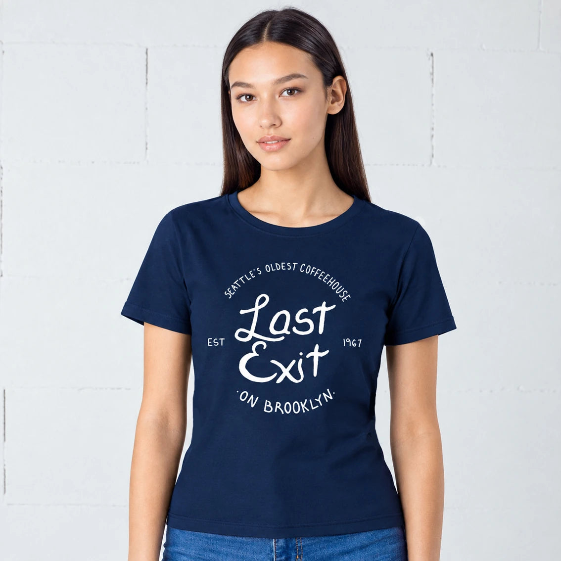 A woman modeling a navy blue short-sleeve t-shirt with a white graphic print that reads 'Last Exit on Brooklyn' and 'Seattle's Oldest Coffeehouse'.