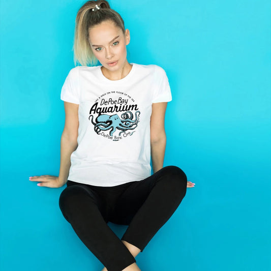 A woman sitting against a bright blue background wearing a white graphic t-shirt with a blue octopus design and the text 'DePoe Aquarium' 